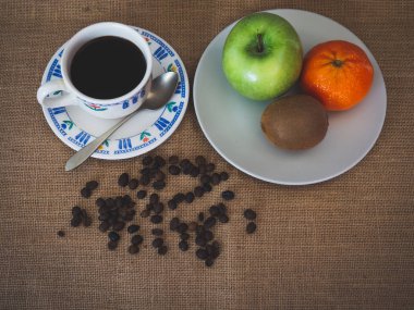 Porselen kahve fincan, kahve çekirdekleri, eski bir kaşık ve bir tabak bir yeşil elma, bir mandalina ve vintage çuval bezi arka plan üzerinde bir kivi ile kahvaltı