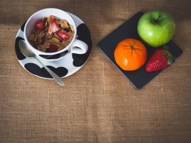 Bir porselen kase tahıl kırmızı meyveler, eski bir kaşık ve bir tabak bir yeşil elma, bir mandalina ve vintage çuval bezi arka plan üzerinde bir kivi ile kahvaltı