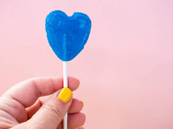 Een vrouw met geel geschilderde nagels met een blauwe lolly in haar hand en pastel roze achtergrond — Stockfoto