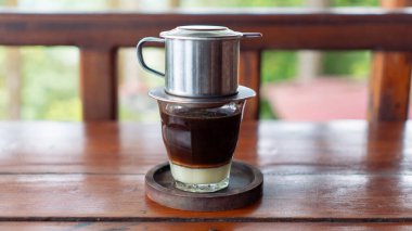 Viyana Kahvesi ya da Vietnam Drip, üzerinde yoğun süt ve metal damlaları olan bir bardakta sunuldu. Köy ahşap bir masaya yerleştirilmiş, zengin ve lezzetli bir içecek demlemeye hazır..
