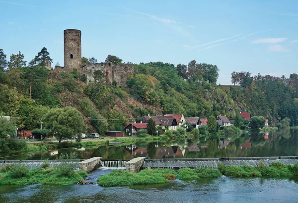 Tower of castle ruins on a hill surrounded by green plants. Village buildings and river with weir are under the castle ruins. Castle ruins Dobronice near Bechyne, Czechia.