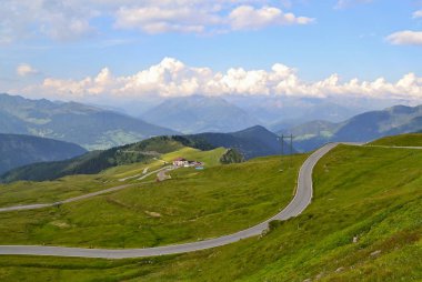 Güzel Alps dağlar ülkede Tyrol. Yol bir bina için yeşil çimenli dağlarda büküm. Orman ve yüksek tepeler arka planda vardır. Bulutlar parlayan beyaz tabakası tepeler ve mavi gökyüzü arasında bulunmaktadır. Tyrol, İtalya