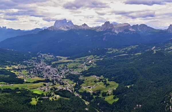 Cortina D Ampezzo aerial picture - city between green forest and mountain peaks. Photo of the city from a mountain peak. City is surrounded by a green forest. High rocky mountains peaks and cloudy sky are over the city. Cortina D Ampezzo, Dolomites,