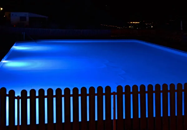 Blue illuminated pool at a night with a wooden black fence in foreground. Lights are shining on the pool, darkness is all around.