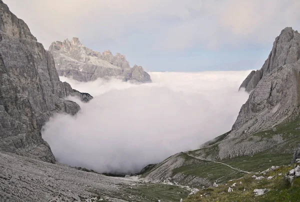 Fog rolling up in a valley between rocks in mountains. High rocky mountains are around, hiking trail goes into the fog. Green grassy places are in the foreground and some clouds are on the sky top. Italy, Italian Alps - Dolomites.