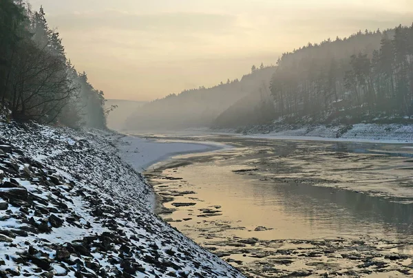 River with steep rocky banks and ice in the water. River in a deep watercourse - a partially drained water dam. The banks are from stones. A forest is around the banks and pieces of ice are in the water. Czechia, Hracholusky dam, december 2018