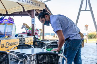 Huelva, İspanya - 4 Temmuz 2020: Yüzü maskeli bir garson Islantilla ticaret merkezindeki barın masasını dezenfektan sıvısıyla temizliyor. Yeni normalde Coronavirus covid-19 'a karşı koruma