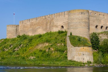 Narva, Estonya - 13 Temmuz 2025: Ivangorod Kalesi 'nin manzarası. Ortaçağ taş duvarları, konik çatılı yuvarlak kuleler, nehir kenarındaki yeşillikler ve açık mavi gökyüzünün altında akan Narva Nehri.