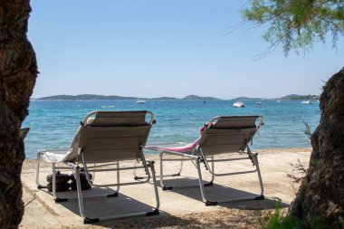 Ağaçların altında havluları olan iki plaj şezlongu kristal mavi denize ve uzak adalara bakan, suda tekneleri olan. Yaz tatili, dinlenme, seyahat, boş zaman, kıyı yaşam tarzı, Akdeniz, tatil ve doğa temaları için ideal.