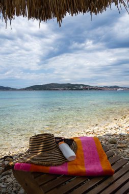 Plaj tatili esasları güneş kremi, güneş kremi, güneş gözlüğü ve renkli havluyu kamış şemsiyenin altında, parlak mavi su ve şehir silueti ile birlikte. Yaz, seyahat, rahatlama, rahatlama ve sağlık konuları için mükemmel..