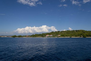 Mavi denizi olan kıyı manzarası, yeşil yamaç, kıyı boyunca evler, ve gökyüzünde dramatik bulutlar. Seyahat, tatil, doğa, sükunet ve manzaralı Akdeniz veya Avrupa gezileri için mükemmel