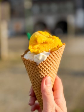İki kepçe dondurmayla dolu waffle külahını tutan bir elin yakın çekimi. Bir kepçe sarı mango ve bir kremalı vanilyalı açık hava bulanıklığına karşı. Görüntü dondurmadan zevk almanın ferahlatıcı zevkini ve neşesini yakalar..