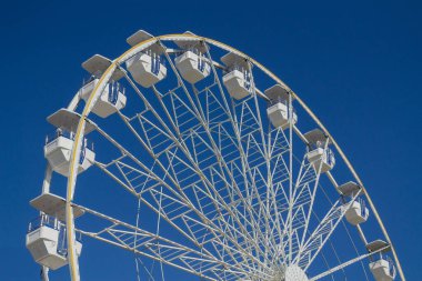 Parlak mavi gökyüzü altında mavi aksanlı kulübeleri olan zarif beyaz dönme dolap, yaz, seyahat, eğlence, aile eğlencesi, turizm ve parklarda ya da şehir manzaralarında açık hava macerası temaları için mükemmel..