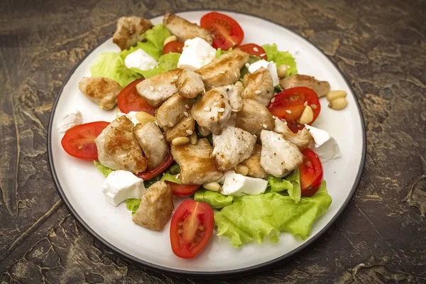 Delicious salad with warm chicken, cherry tomatoes and soft brynza cheese, decorated with pine nuts. dressing with olive oil. Dish on old cutting board