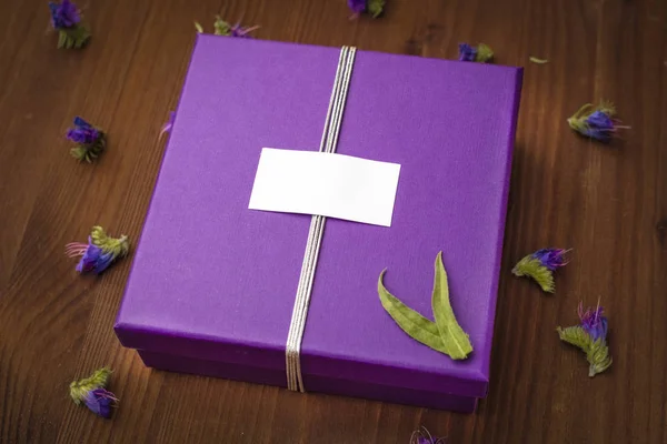 Violet gift box with blank tag on a wooden table with small flowers. Womans day, 8 march, wedding, dating, love concept. Top view