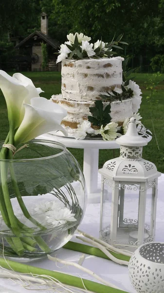Kutlamalar için beyaz süsleme masası - iki katlı pasta, beyaz fener ve yuvarlak cam vazoda zambak
