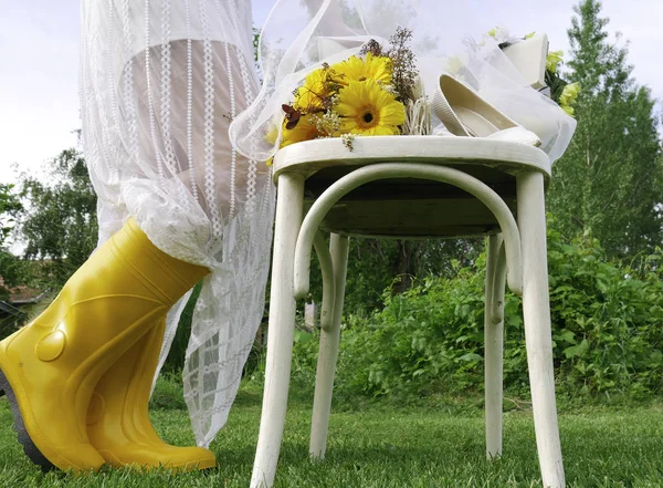 黄色のガンブーツと白い春のドレスの女性は木製の椅子と黄色のガーベラデイジーの花束の横にある — ストック写真