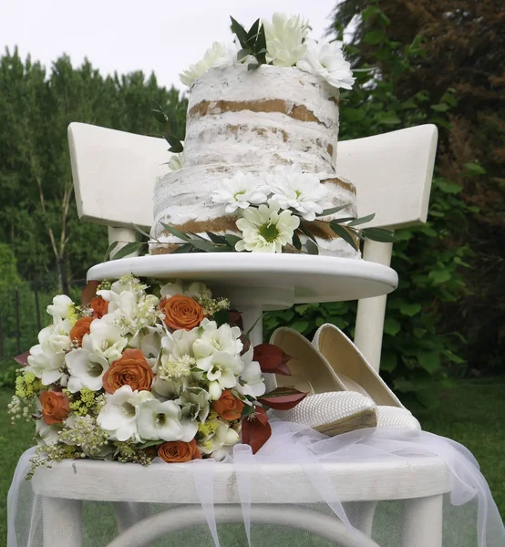 Two-tiered cake, spring bouquet and white bride\'s shoes on a white wooden chair - outside