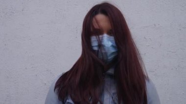 Rüzgar saçlarını taşırken dışarıda beyaz bir duvarın önünde cerrahi maske takan bir kadının portresi..