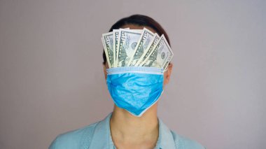 Kadın doktor ya da hemşire, tıbbi maskesinden sarkan 50 dolarlık banknotlarla gözlerini kapatıyor..