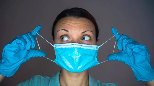 A female doctor or nurse with rubber gloves on her hands puts a blue mask on her face.