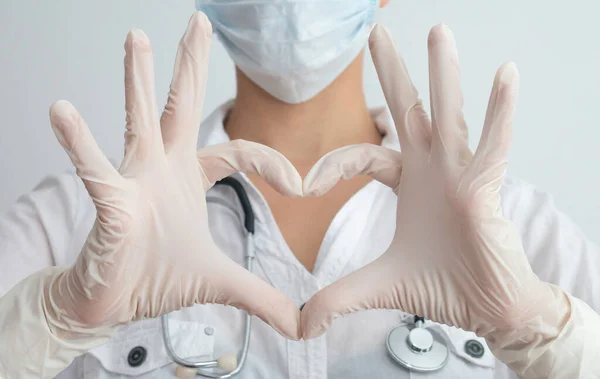 Female doctor with mask, gloves and stethoscope. Woman with a medical mask and hands in latex glove shows the symbol of the heart. Doctor for the heart.