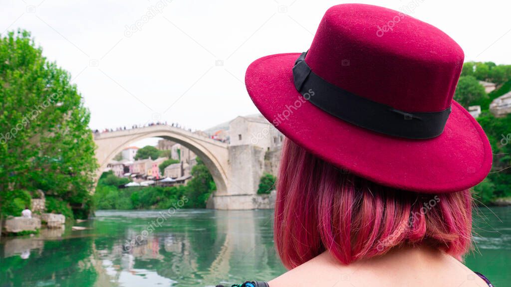 Photographed from the back of a woman's head - A woman with pink hair and a hat looks at the Old bridge over the river Neretva in Mostar,  Bosnia and Herzegovina.
