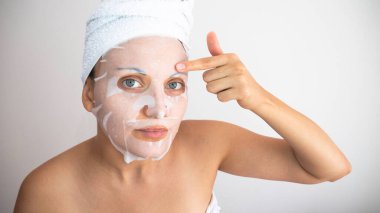 Genç bir kadın beyaz arka plana beyaz kozmetik kumaş yüz maskesi takıyor. Yüz soyma maskesi. Güzellik merkezi. 