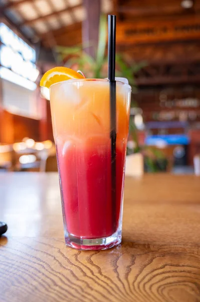 Tropikal kokteyl, katmanlı kırmızı ve turuncu içecek ahşap masada uzun bardakta.