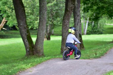 Güvenlik kaskı takan küçük çocuk kaldırımlı park yolunda yeşil çimlerle çevrili kırmızı dengeli bisiklete biniyor. Çocukluk macerası ve sağlıklı yaşam tarzı..