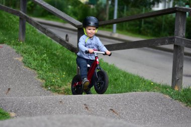 Kask takan genç çocuk yüksek kaldırımlı açık hava pompa pistinde kırmızı dengeli bisiklete biniyor. Yeşil park ortamında heyecan verici çocukluk sporları ve aktif oyun zamanı.