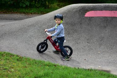 Kask takan genç çocuk yüksek kaldırımlı açık hava pompa pistinde kırmızı dengeli bisiklete biniyor. Yeşil park ortamında heyecan verici çocukluk sporları ve aktif oyun zamanı.