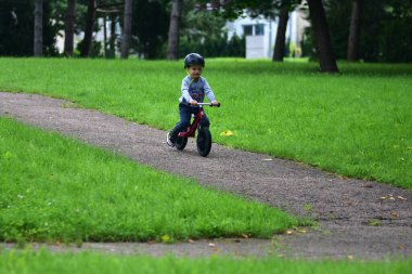Güvenlik kaskı takan küçük çocuk kaldırımlı park yolunda yeşil çimlerle çevrili kırmızı dengeli bisiklete biniyor. Çocukluk macerası ve sağlıklı yaşam tarzı..