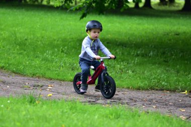 Güvenlik kaskı takan küçük çocuk kaldırımlı park yolunda yeşil çimlerle çevrili kırmızı dengeli bisiklete biniyor. Çocukluk macerası ve sağlıklı yaşam tarzı..
