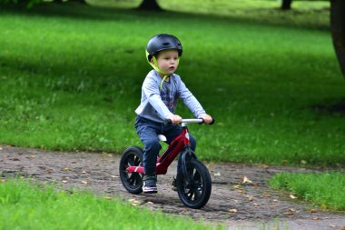 Güvenlik kaskı takan küçük çocuk kaldırımlı park yolunda yeşil çimlerle çevrili kırmızı dengeli bisiklete biniyor. Çocukluk macerası ve sağlıklı yaşam tarzı..