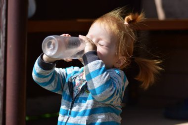 Kızıl saçlı küçük kız gün ışığında şeffaf şişeden su içiyor. Güzel bir çocukluk anı, sağlıklı su kaybı ve basit bir aile hayatı..