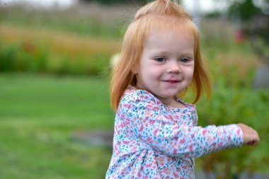 Close-up portrait of happy red-haired toddler in floral shirt outdoors. Child smiling with innocent expression in natural green garden background.