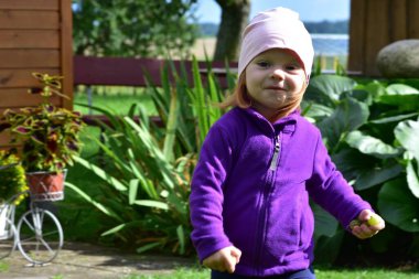 Happy little girl in purple jacket and pink hat runs outside near wooden house, holding small fruits. Joyful childhood playtime in countryside garden with natural sunlight.