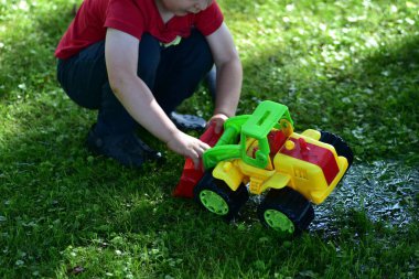 Child crouching and playing with colorful plastic toy tractor on wet grass with water, summer outdoor play. Childhood imagination and fun activity.