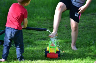 Children playing with garden hose and toy tractor, splashing water on grass in summer. Fun outdoor childhood activity and family leisure.