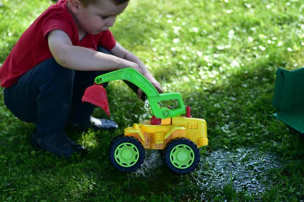 Child crouching and playing with colorful plastic toy tractor on wet grass with water, summer outdoor play. Childhood imagination and fun activity.