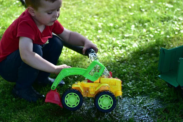 Child crouching and playing with colorful plastic toy tractor on wet grass with water, summer outdoor play. Childhood imagination and fun activity.