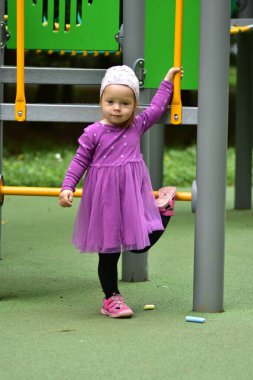 Cute toddler girl in purple dress and hat leaning on playground structure. Child holds yellow handle and poses casually while enjoying outdoor playtime in park.