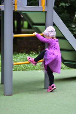 Little girl in purple dress climbing up yellow ladder at playground. Toddler enjoying outdoor activity, developing strength and coordination during playful park day.