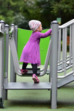 Mor elbiseli küçük bir kız çocuğu ve modern oyun alanının merdivenlerine tırmanıyor. Dışarıda oynarken denge ve güç uygulayan çocuk.