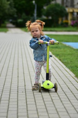 Red-haired toddler girl in denim jacket and leggings riding green scooter on paved sidewalk. Child practicing balance and coordination while enjoying outdoor play.