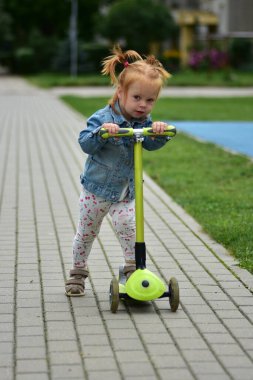 Red-haired toddler girl in denim jacket and leggings riding green scooter on paved sidewalk. Child practicing balance and coordination while enjoying outdoor play.