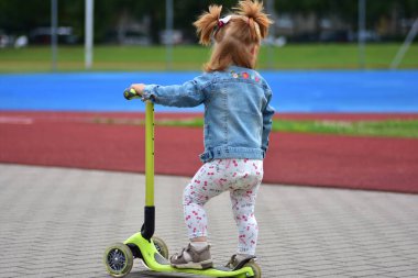 Kot ceketli at kuyruklu küçük kız mavi koşu pisti ve kırmızı atletizm sahasının yanında scooter sürüyor. Çocuk dışarıda egzersiz ve eğlenceli yaz eğlencesiyle eğleniyor..