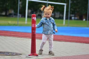 Açık hava spor sahasında kırmızı koninin yanında duran kot ceketli ve baskılı taytlı kızıl saçlı küçük kız. Çocuk merakla çevreyi keşfediyor.