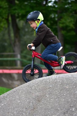 Smiling child in helmet riding red balance bike on top of pump track hill. Little boy outdoors in autumn park, enjoying active sport, balance practice and playful cycling.